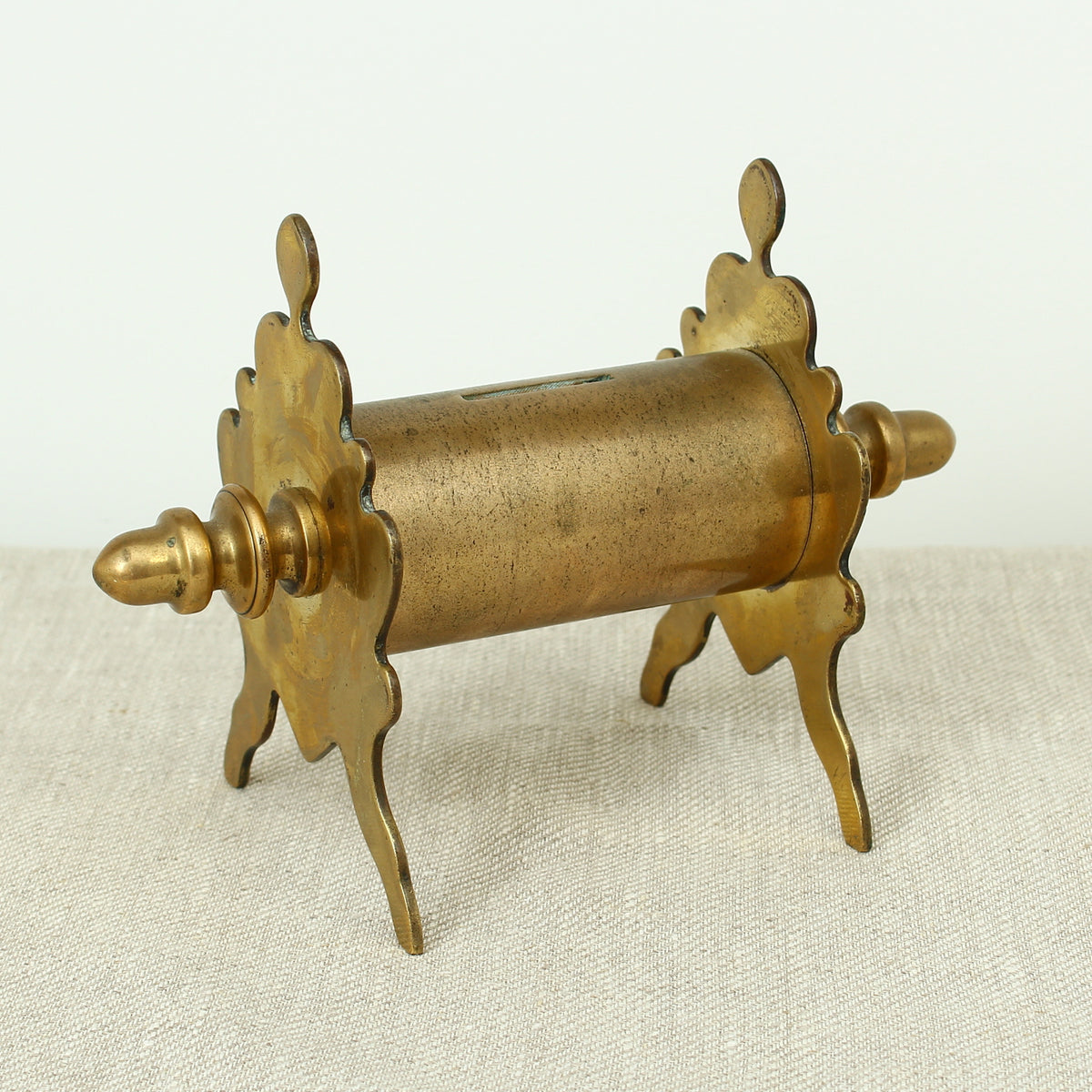 A late 19th century or early 20th century brass money box/bank, cylindrical central section with single money slot, shaped end pieces forming a combination pen rest, threaded finials to each end, 13.5cm high, 20cm wide