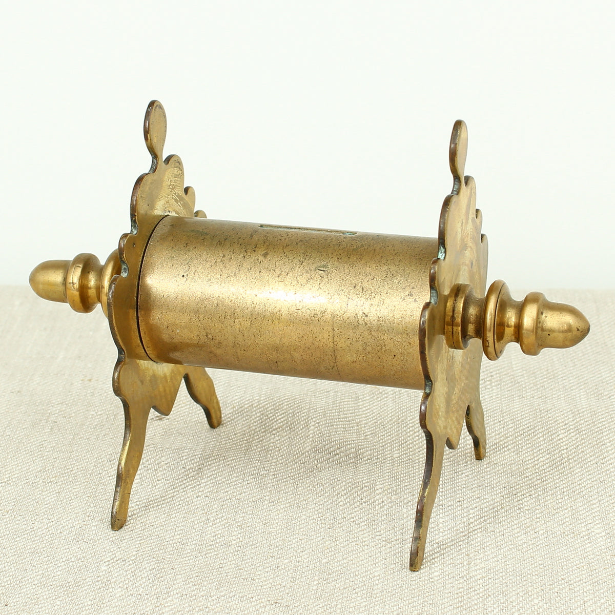 A late 19th century or early 20th century brass money box/bank, cylindrical central section with single money slot, shaped end pieces forming a combination pen rest, threaded finials to each end, 13.5cm high, 20cm wide
