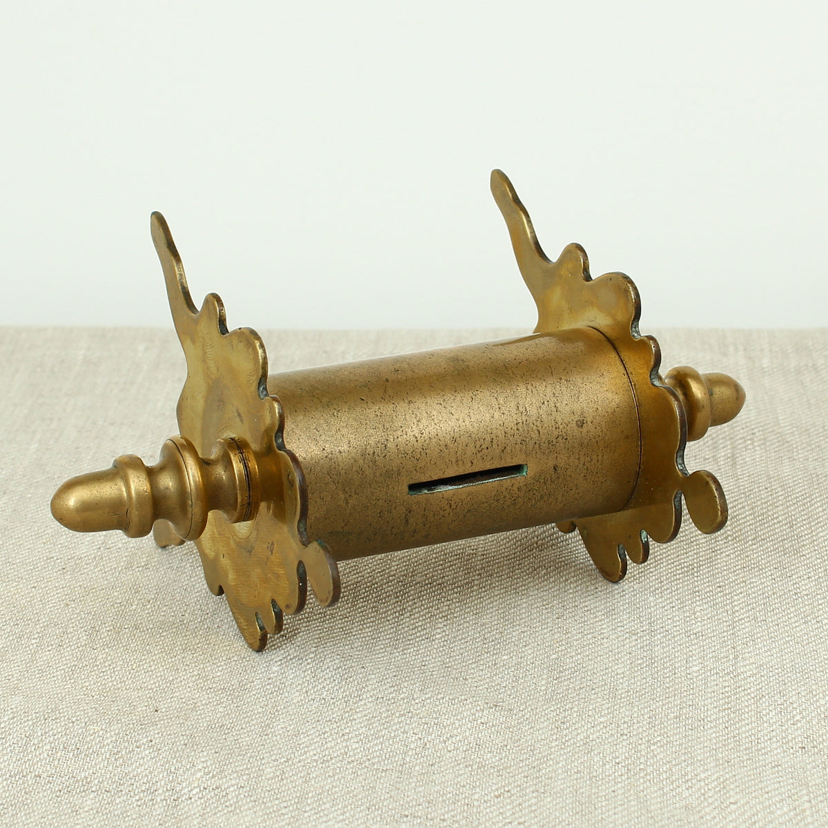 A late 19th century or early 20th century brass money box/bank, cylindrical central section with single money slot, shaped end pieces forming a combination pen rest, threaded finials to each end, 13.5cm high, 20cm wide