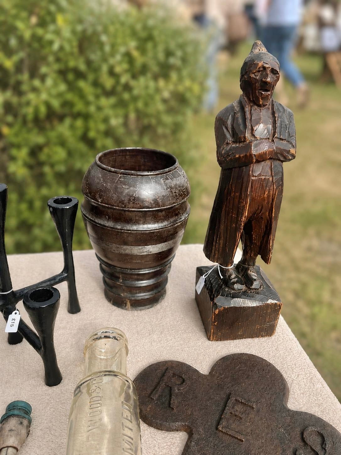 A collection of vintage and antique objects displayed outdoors on a table, including a carved wooden figurine of a standing man, a dark metal pot, black metal pipe holders, a clear glass bottle, and a dark cast-iron piece engraved with 'RES'.